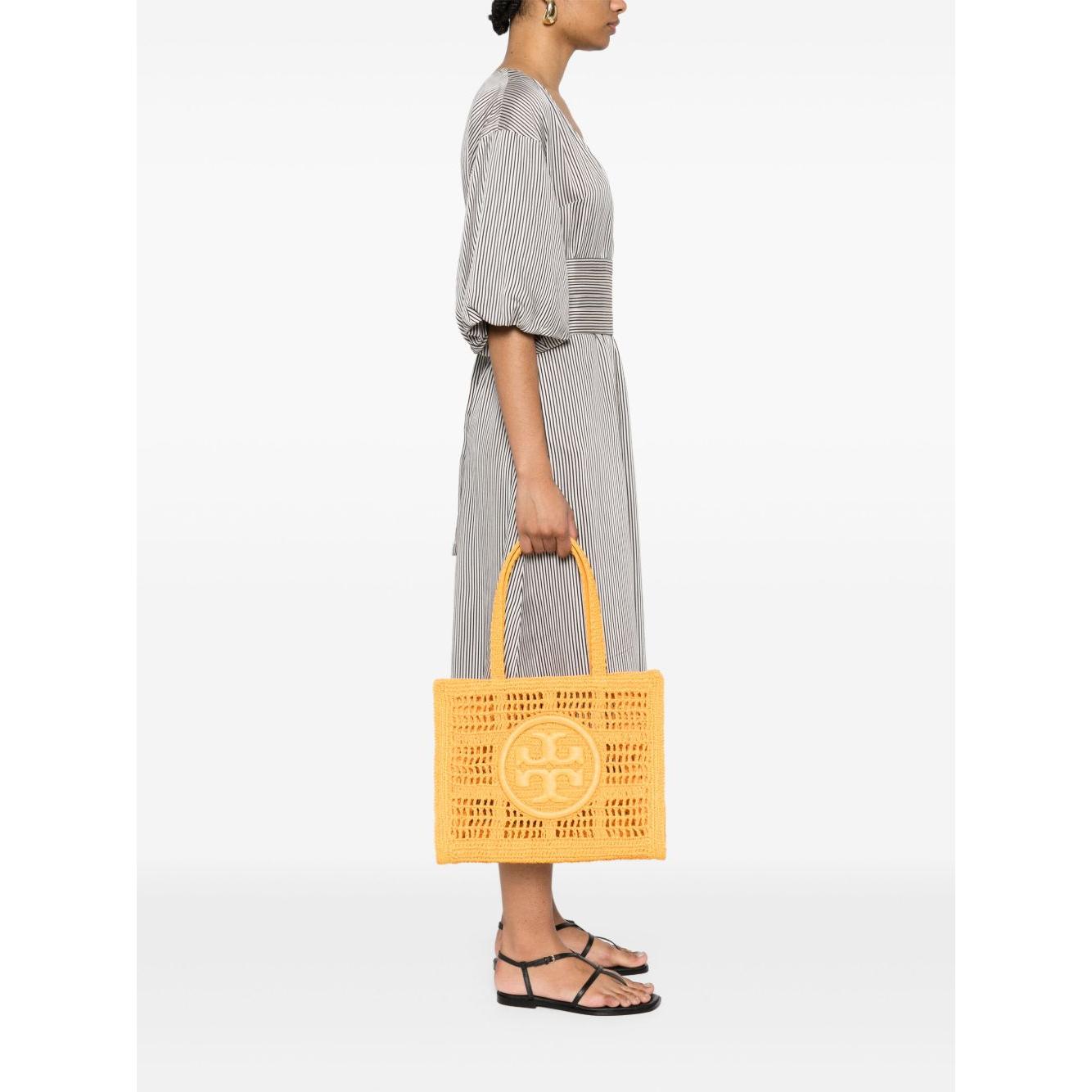 Woman holding Tory Burch Ella Small carrot orange raffia tote bag with signature Double T motif and top handles.