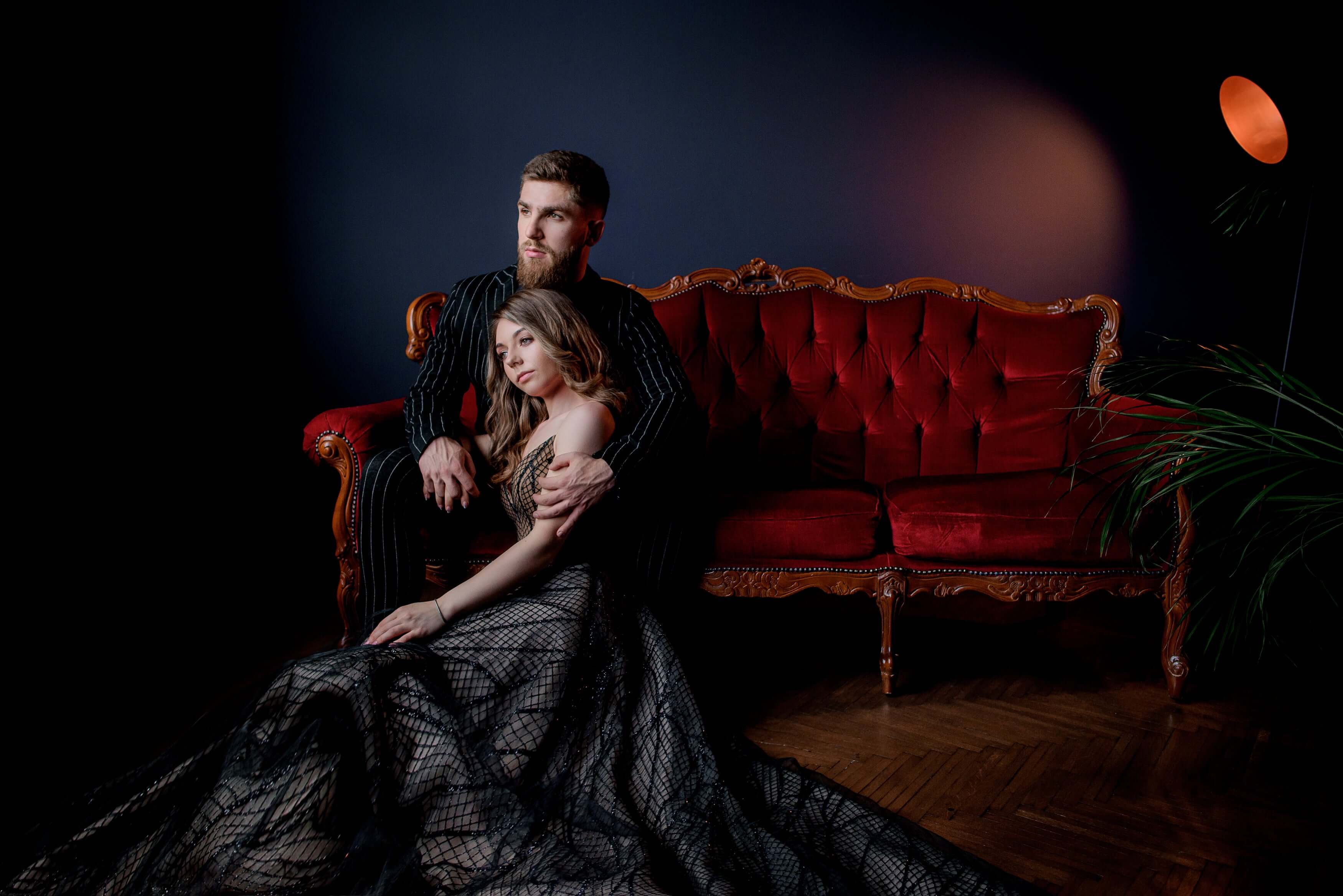 Elegant couple on a vintage red velvet sofa, woman in a black gown posing with a dark backdrop and warm lighting ambiance.